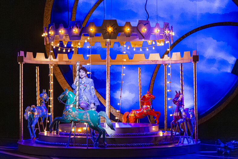 Een toneelvoorstelling met een draaimolen, verlicht door lichten, met een performer in een kleurrijk kostuum.