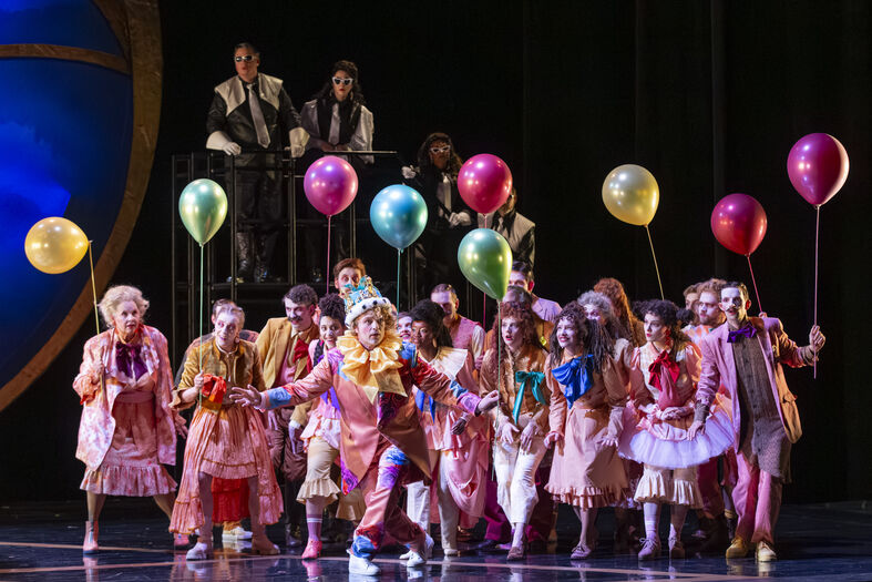Een groep acteurs in kleurrijke kostuums, met ballonnen, op het podium van een theater. Achtergrond met toeschouwers.