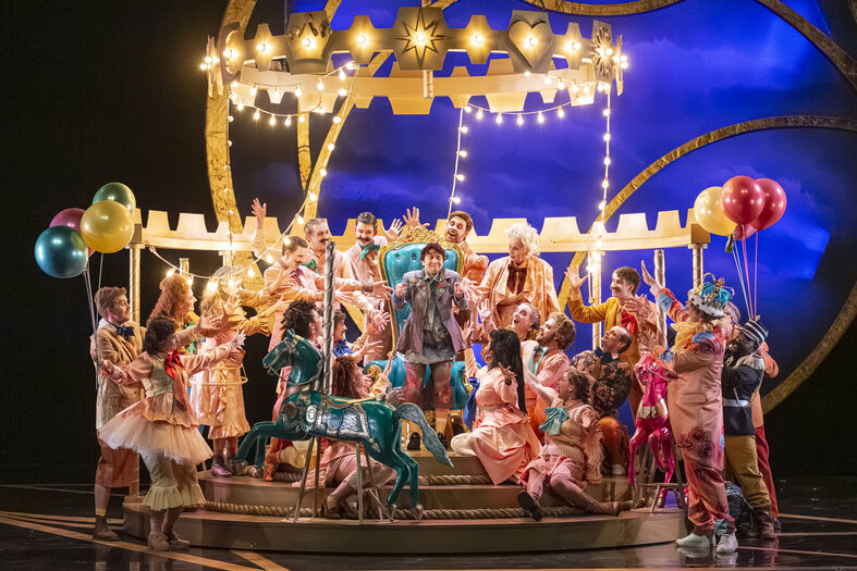 Een kleurrijke theaterproductie met dansers en acteurs op een festieve set met ballonnen en verlichting.
