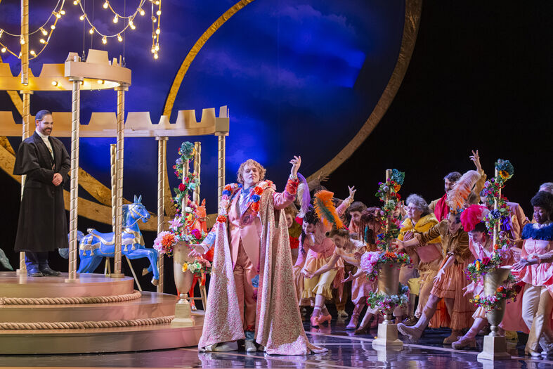 Een theatrale scène met acteurs in kleurrijke kostuums, een verhoogd podium en een publiek dat enthousiast reageert.