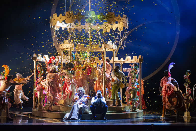 Theatrale voorstelling met kleurrijke kostuums, confetti en een decor van een grote kroonluchter, met acteurs op het podium.