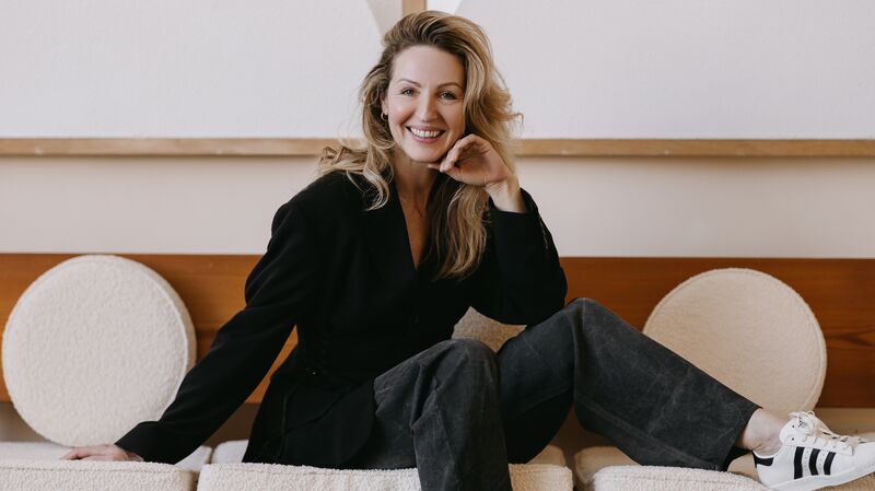 Debby Gerritsen zit ontspannen op een bank met lichte ronde kussens, terwijl een ander op de leuning leunt in een modern interieur met geometrisch reliëf aan de wand.