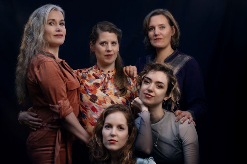 Groep van vijf vrouwen poseren samen met verschillende gezichtsuitdrukkingen en kledingstijlen tegen een donkere achtergrond.