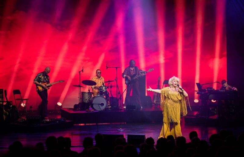 Een muzikante zingt op het podium met een band, omgeven door kleurrijke lichteffecten.