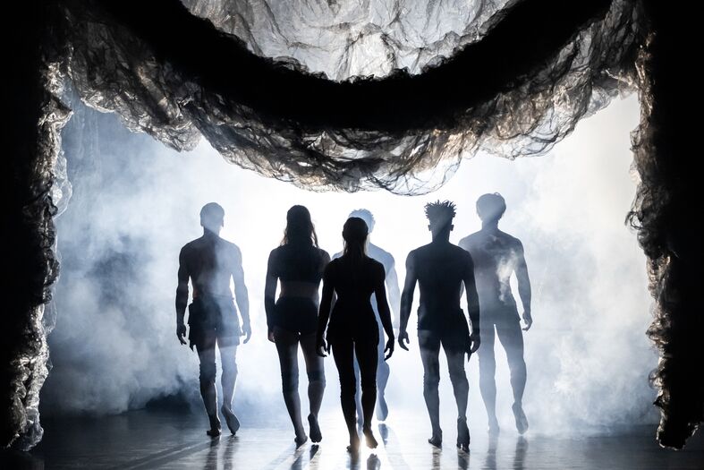 Vier silhouetten van dansers staan op het podium, omgeven door mist en dramatische belichting.