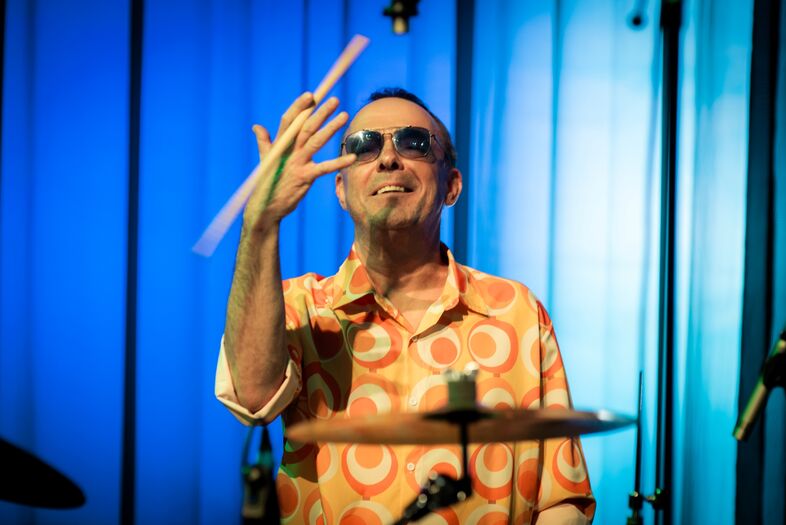 Drummer in een kleurrijk shirt speelt op een podium, met een energieke uitstraling en zonnebril.
