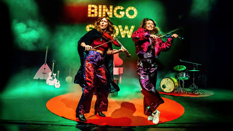 Twee muzikanten in kleurrijke outfits dansen en spelen viool op een podium met een kleurrijke achtergrond en instrumenten.