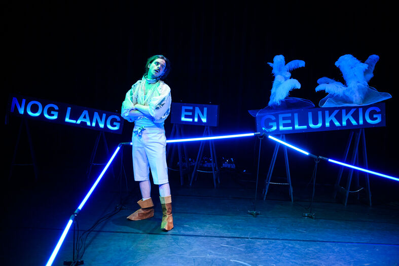 Een performer in witte kleding staat met een zelfverzekerde houding tussen twee neonborden met de tekst "nog lang en gelukkig."