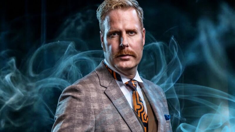 Man in een geruit kostuum met een snor, staand tegen een achtergrond van blauwe rook.