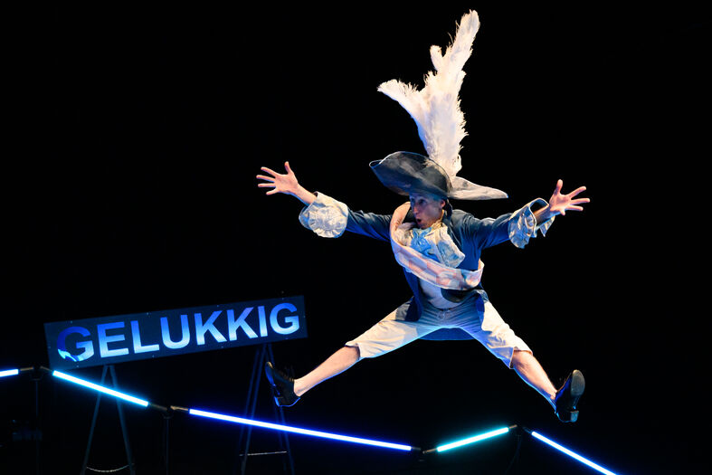 Een acrobaat in een kostuum springt met uitgestrekte armen op een podium met de tekst "GELUKKIG" op de achtergrond.