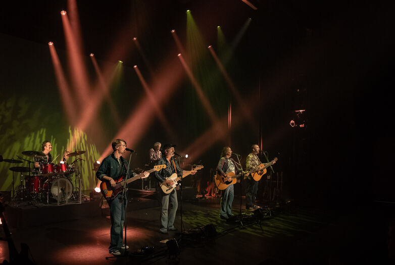 Zes muzikanten spelen op een podium met kleurrijke verlichting en een band in de achtergrond.