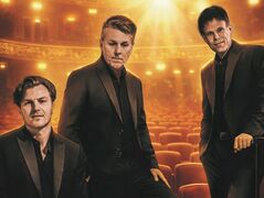 De 3JS in donkere pakken poseren in een warm verlicht theater met rijen rode stoelen en gouden lichtaccenten.