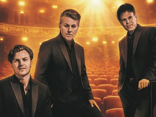 De 3JS in donkere pakken poseren in een warm verlicht theater met rijen rode stoelen en gouden lichtaccenten.