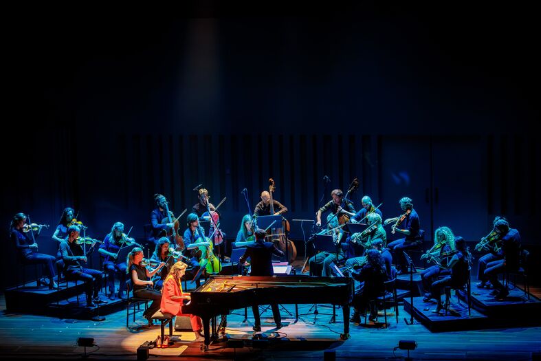 Een pianist en een orkest spelen samen op een podium, omringd door blauwe en groene lichten.