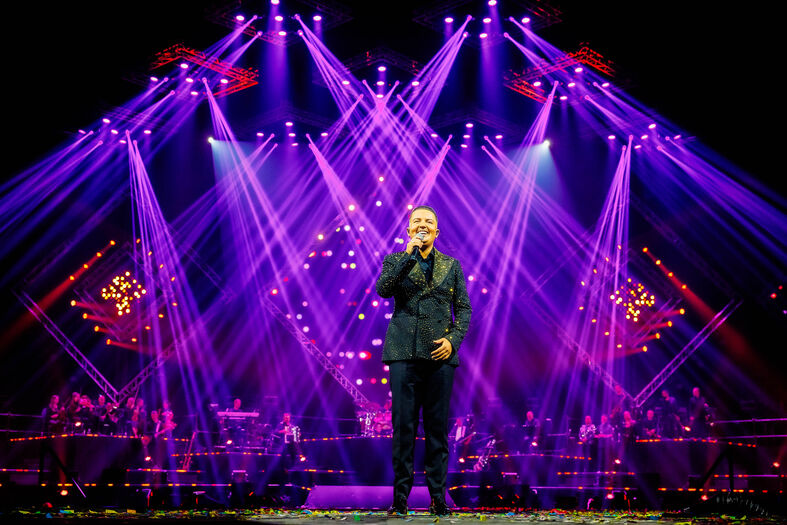 Marco Schuitmaker staat centraal onder paarse lichtstralen terwijl band en dansers op de achtergrond optreden.