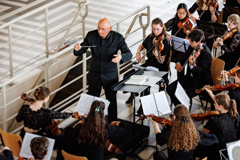 Alt-tekst: Een dirigent leidt een orkest van jonge muzikanten in een concertzaal, met violen en bladmuziek zichtbaar.