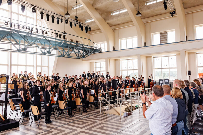 Een groep muzikanten en zangers op een podium in een grote zaal, terwijl het publiek applaudisseert.