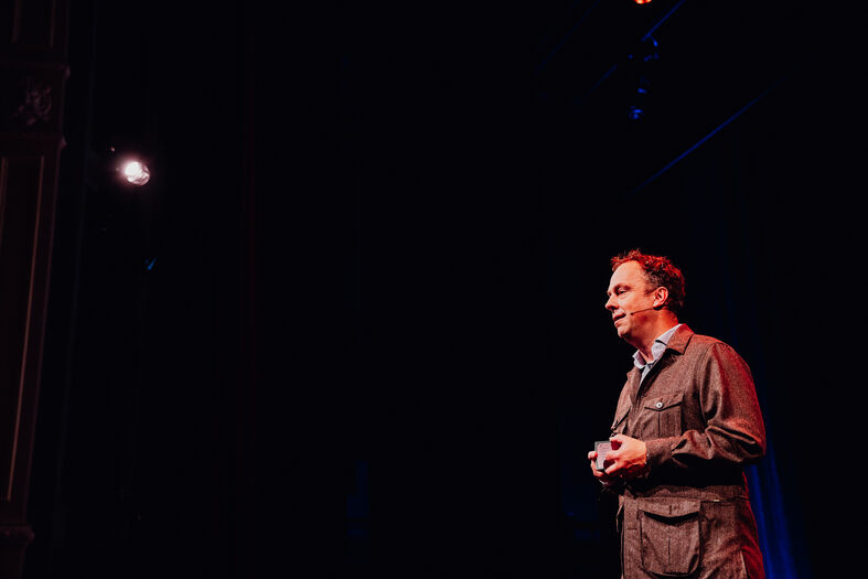 Man in een donker theater spreekt op een podium, verlicht door een enkele lichtbron.