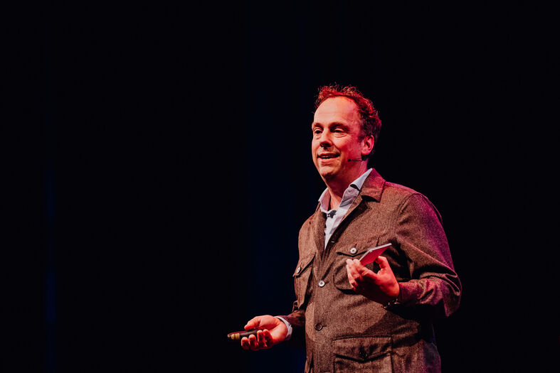 Een presentator in een donker theater, die met een microfoon en een object in zijn hand spreekt.