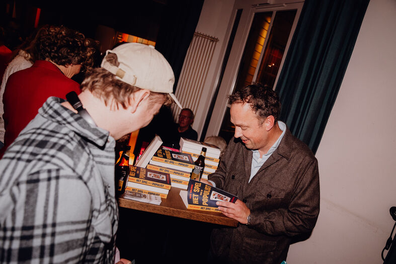 Twee mannen in gesprek bij een tafel met stapels boeken in een sfeervolle omgeving.