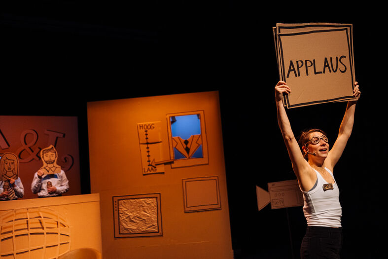 Een performer houdt een bord met de tekst "APPLAUS" omhoog op een podium met theatrale achtergronden.