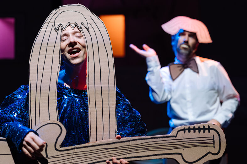 Twee performers op het podium, één met een grote kartonnen gitaar en de ander met een groot, grappig hoofddeksel.