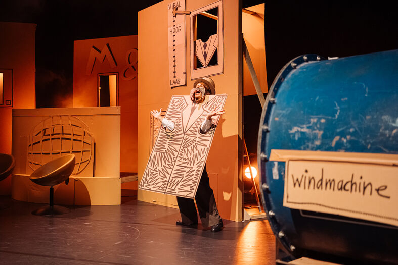 Persoon gekleed in een groot kartonnen bord, poseert op een theaterpodium met decor en een blauwe windmachine.