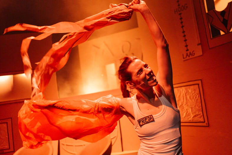 Danseres in een lichte outfit, die een oranje doek in de lucht zwaait, met warme verlichting op de achtergrond.