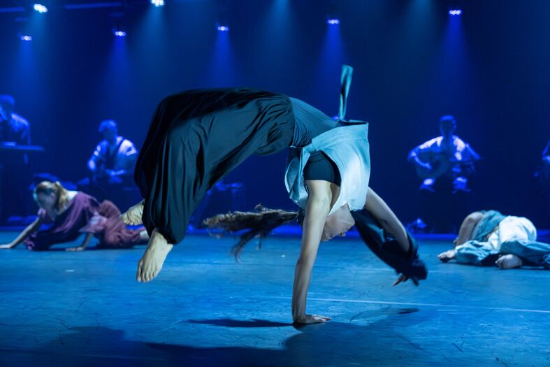 Danseres maakt een acrobatische beweging op een podium, omringd door andere performers in een blauw verlichte setting.
