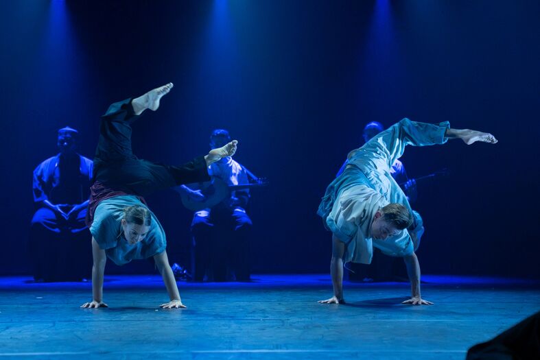 Twee dansers voeren een spectaculaire handstand uit op een podium met blauwe verlichting terwijl muzikanten op de achtergrond spelen.