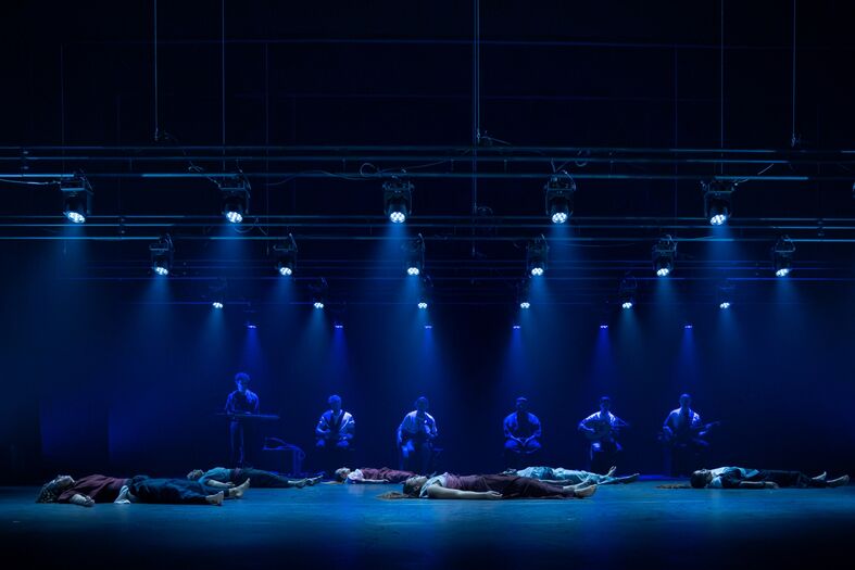 Groep dansers ligt op de vloer in een donkere ruimte, verlicht door blauwe spots.