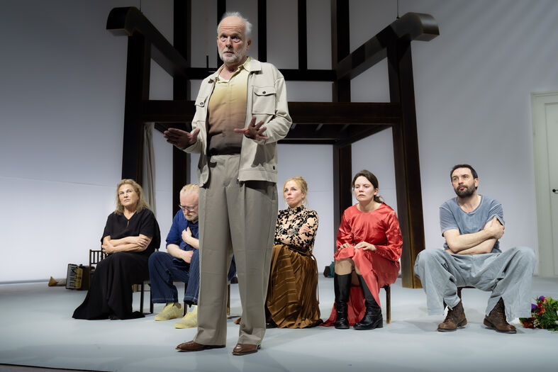 Staande performer spreekt terwijl anderen zittend luisteren onder een grote houten stoel.