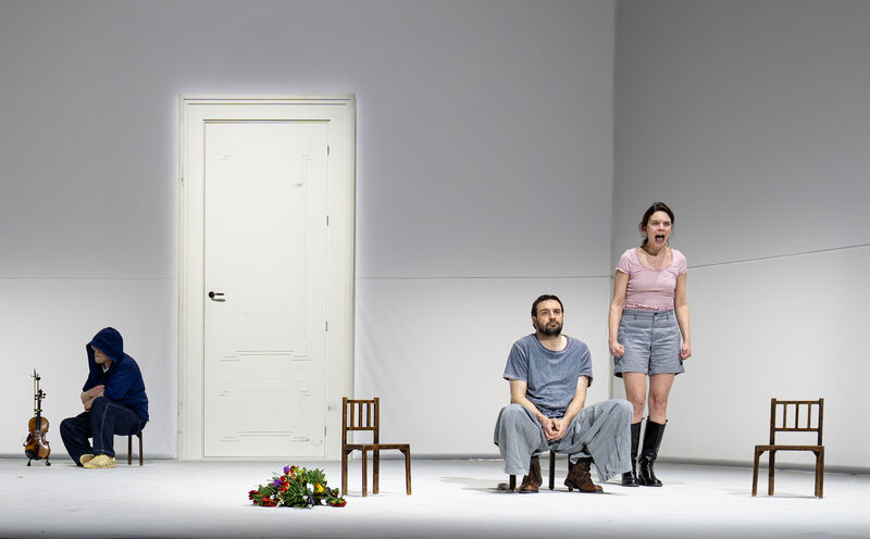 Drie performers op leeg toneel bij witte deur en verspreide stoelen, één zittend met viool naast zich.