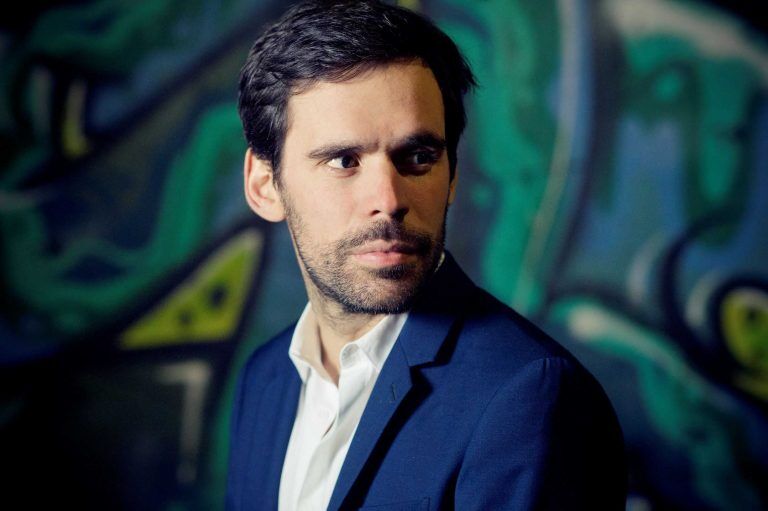 Portret van een man in een blazer met een neutrale uitdrukking, staand voor een kleurrijke graffiti-achtergrond.