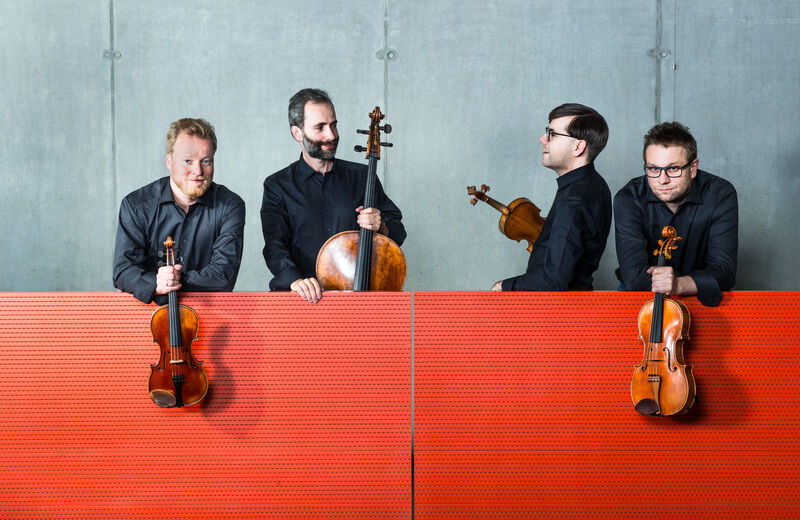 Vier musici in zwarte kleding poseren met strijkinstrumenten voor een rode achtergrond.