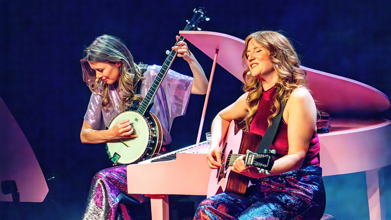 Twee muzikanten spelen op een podium; één met een banjo en de ander met een gitaar, naast een roze piano.