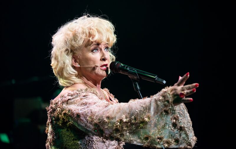 Een vrouwelijke zangeres met blonde krullend haar en een glinsterend kostuum, zingt in een microfoon op het podium.