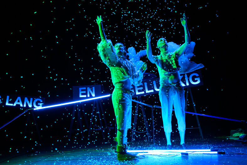 Twee performers in kleurrijke kostuums dansen op een podium met lichteffecten en neerdalende slingers.