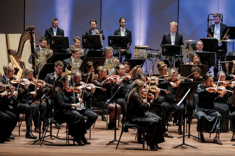 Orkest met strijkers, blazers en slagwerk speelt gezamenlijk op een verlicht podium.