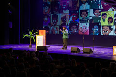 Een spreker op een podium met afbeeldingen op de achtergrond, voor een publiek in een donkere zaal.