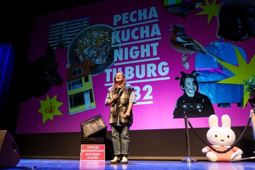Een spreker op het podium tijdens Pecha Kucha Night in Tilburg 2022, met kleurrijke achtergrond graphics.