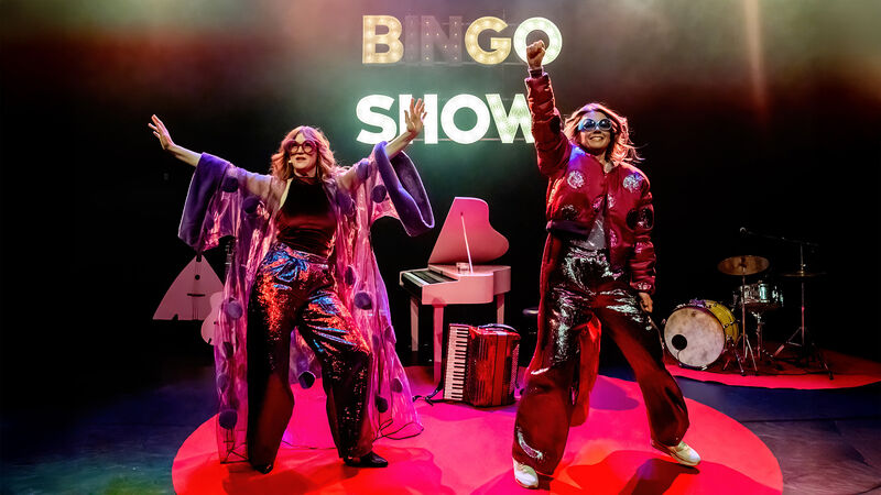 Twee performers in kleurrijke kostuums staan op een podium met de tekst "Bingo Show" op de achtergrond.