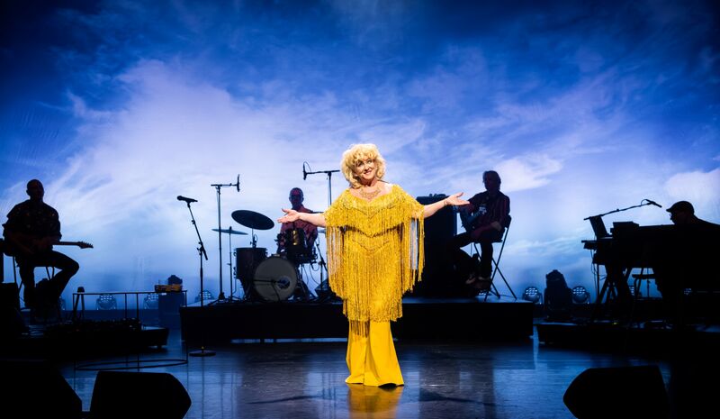 Een artiest in een gele outfit zingt op een podium met een band op de achtergrond, tegen een blauwe luchtachtige achtergrond.