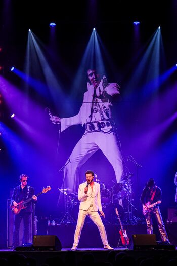 Zanger in witte outfit performt op het podium voor een grote achtergrondbeeld van Elvis Presley, verlichte sfeer.