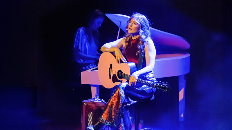 Zangeres met gitaar op een podium, terwijl een pianist op de achtergrond speelt, met sfeervolle belichting.