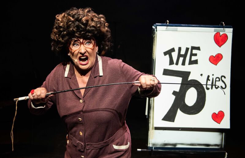 Een vrouwelijke artiest in kostuum staat op het podium met een bord waarop "The 70ties" staat, terwijl ze een interactief optreden geeft.