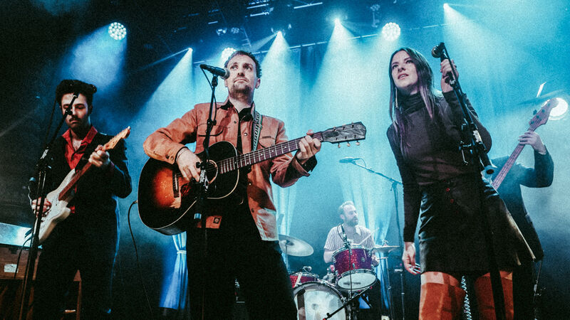 Muzikanten spelen op fel verlicht podium met gitaren, zang en drumstel.