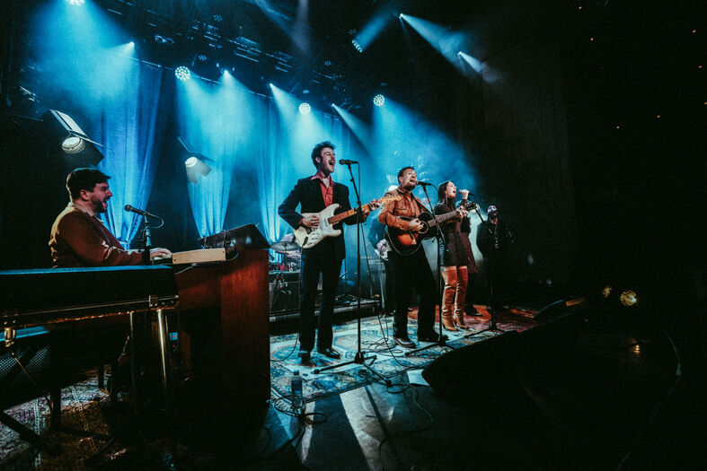 Muzikanten spelen op donker podium met felblauwe lichten en instrumenten.