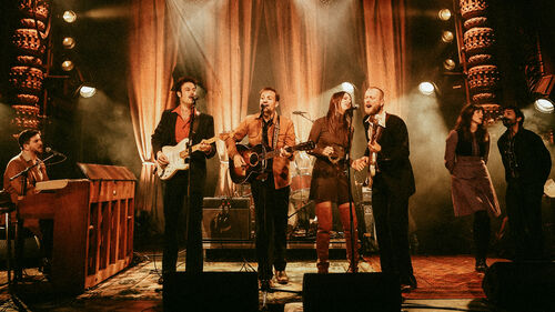 Groep muzikanten speelt op warm verlicht podium met gitaren, piano en zang