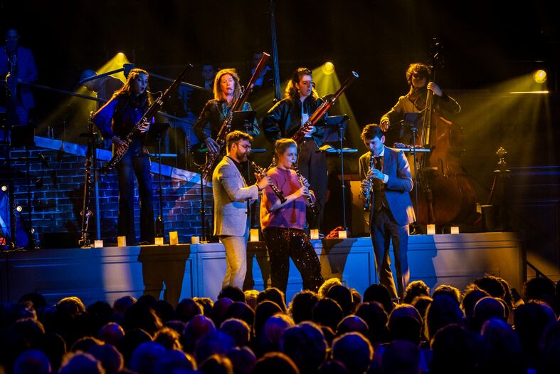 Een live muziekoptreden met een band op het podium, verlicht door kleurrijke verlichting, terwijl het publiek kijkt.